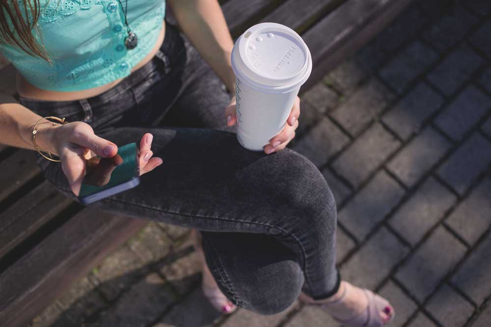 young woman with coffee in hand using self service on mobile phone